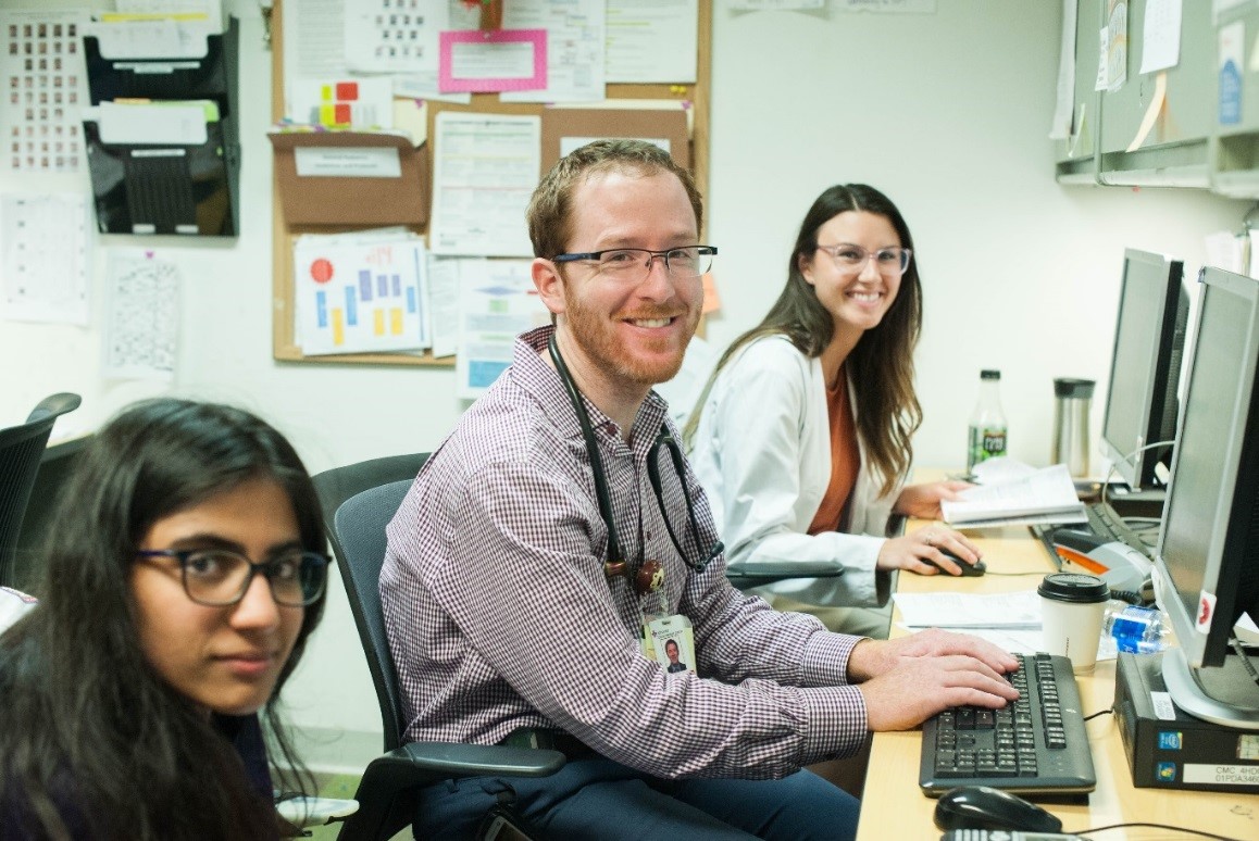 Three doctors look at the camera while they work on desktop computers