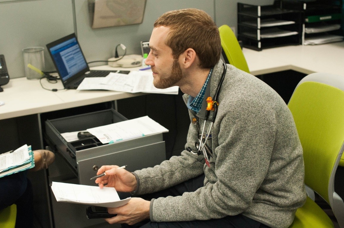 A doctor talks to another doctor while sitting on a chair and taking notes with a pen and notebook